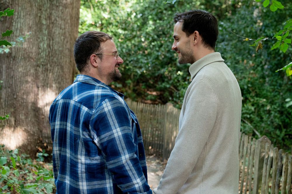 Two men facing each other in conversation outdoors, one in plaid shirt, other in cream sweater, surrounded by trees.