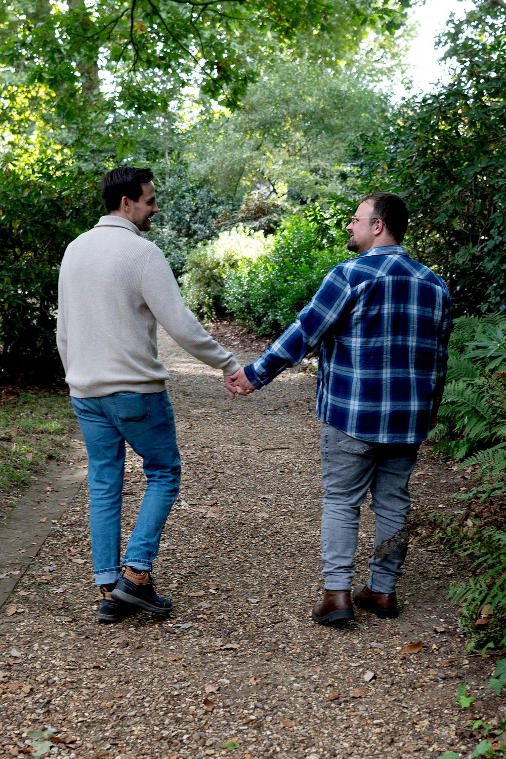 Two men holding hands while walking together on a garden path surrounded by lush green trees and shrubs.