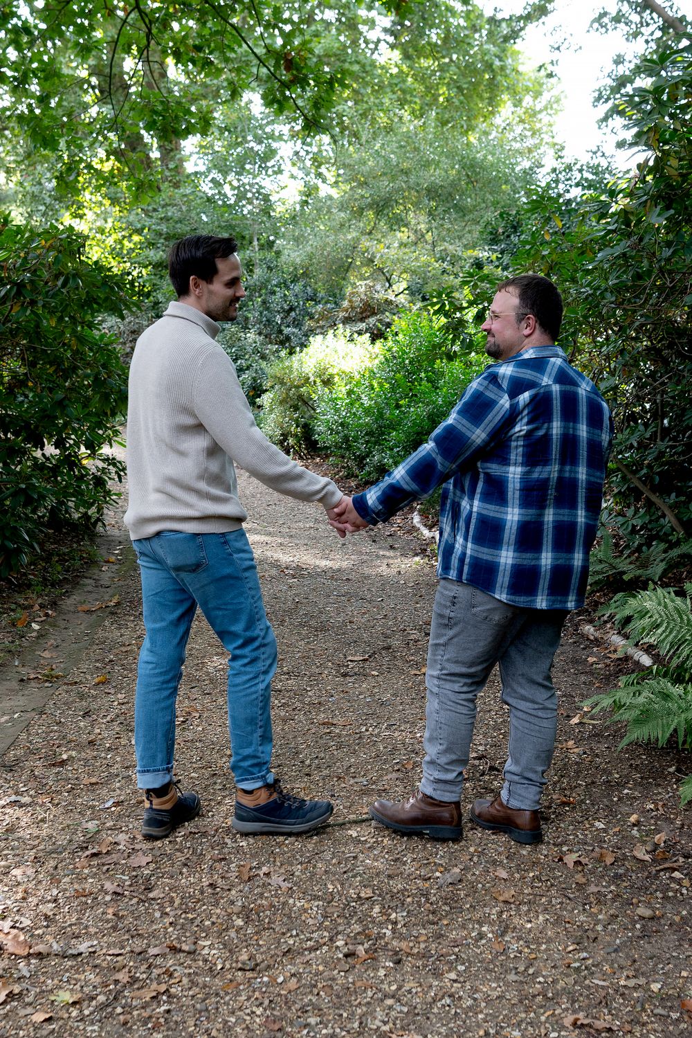 Two men holding hands and smiling at each other on a garden path surrounded by lush green trees and shrubs.