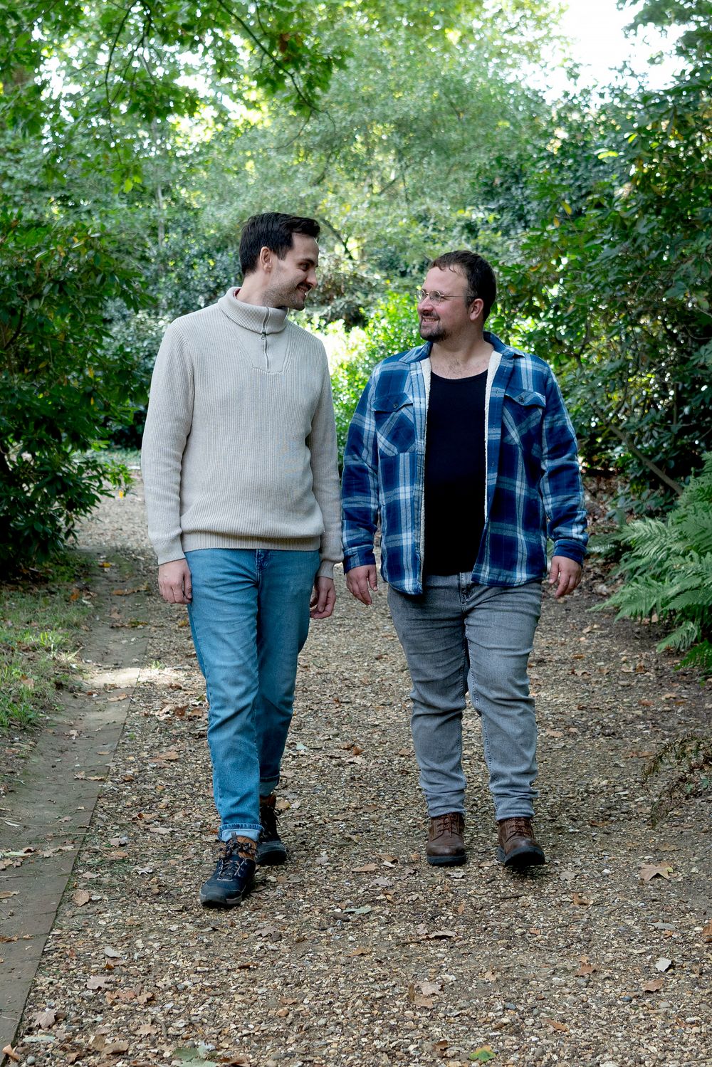 Two men walking together on a gravel garden path surrounded by lush green trees, smiling at each other.