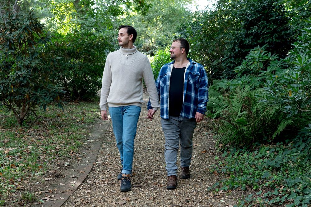 Two men holding hands walk together on a garden path surrounded by lush green trees and shrubs.