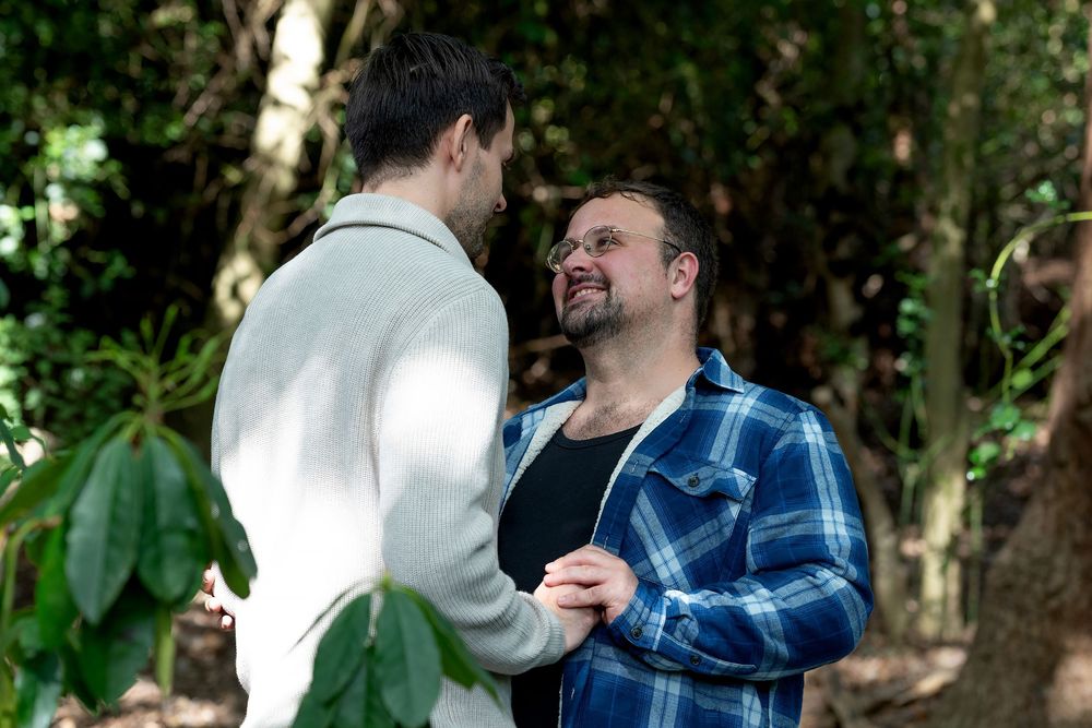 Two men holding hands and smiling at each other in a lush green forest setting.