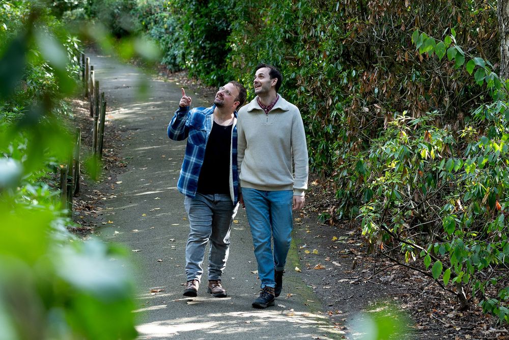 Two men walking along a lush green tree-lined path, one pointing upward while the other looks on curiously.
