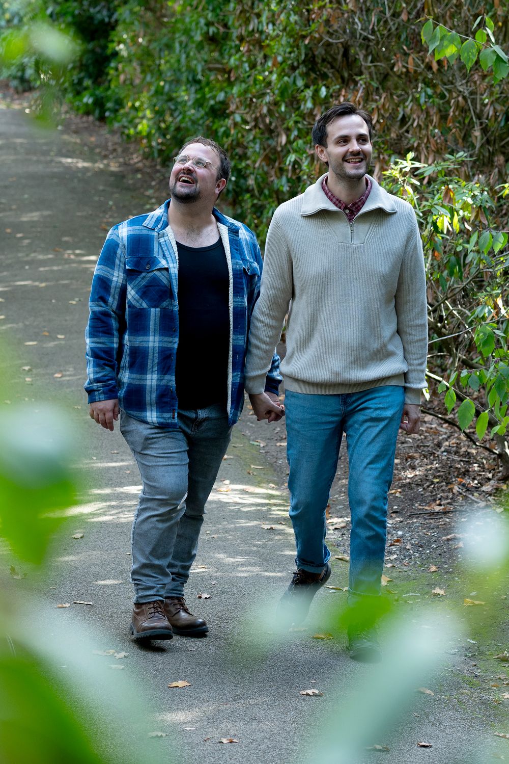 Two men holding hands and smiling while walking together on a tree-lined path outdoors.
