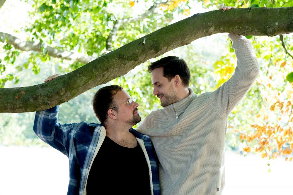 Two men share a tender moment under a tree branch outdoors, surrounded by lush green foliage.