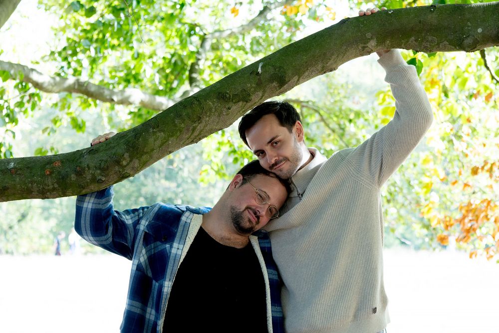Two men pose affectionately under a tree branch outdoors, surrounded by lush green foliage in a park setting.