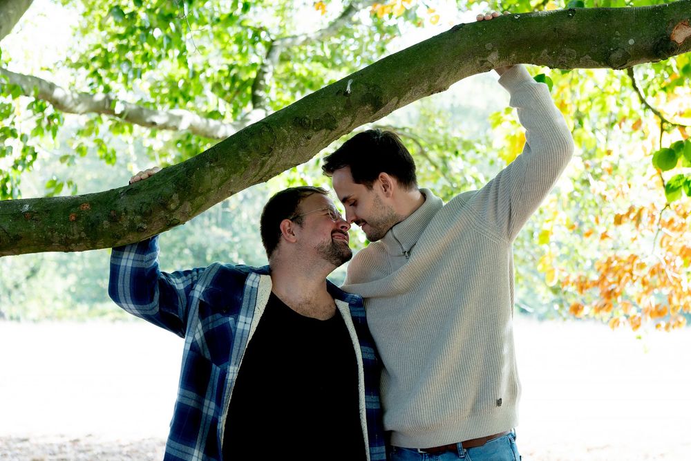Two men share a romantic moment under a tree with lush green leaves in an outdoor park setting.