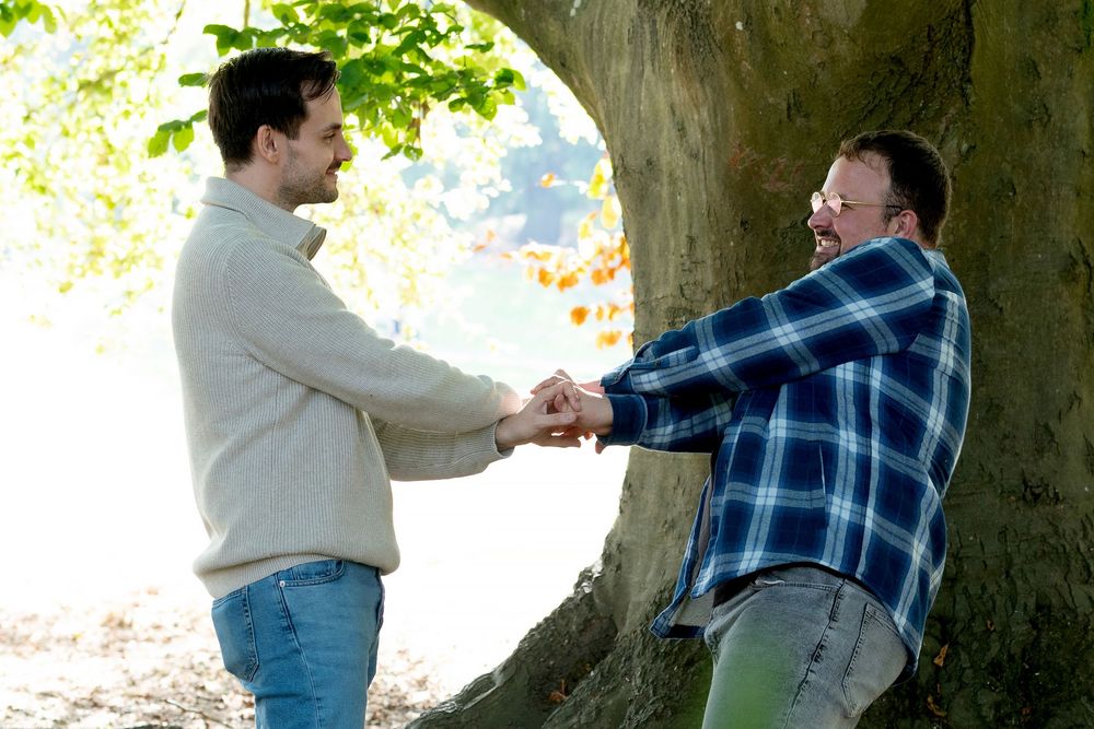Two men holding hands and smiling at each other outdoors near a large tree in a sunny park setting.