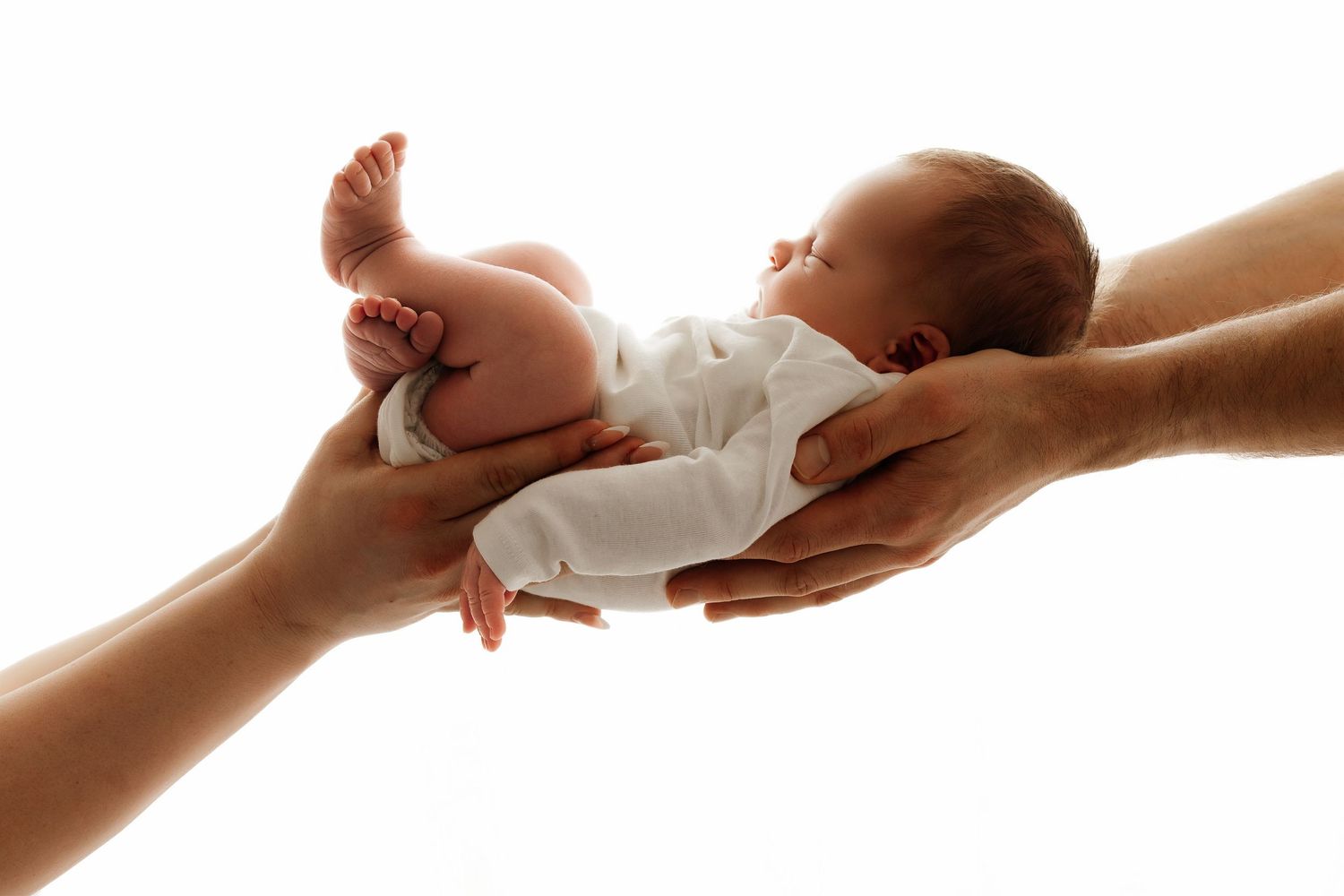 Two pairs of adult hands gently cradling a peaceful newborn baby dressed in white against a white background.