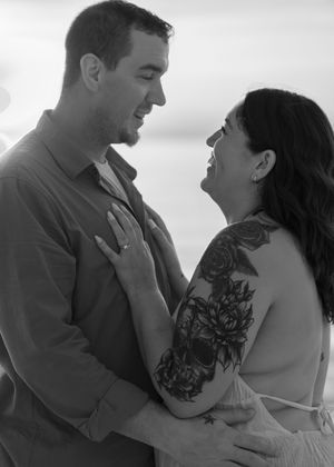 Black and white photo of a couple embracing and smiling at each other, woman with floral tattoo on arm.