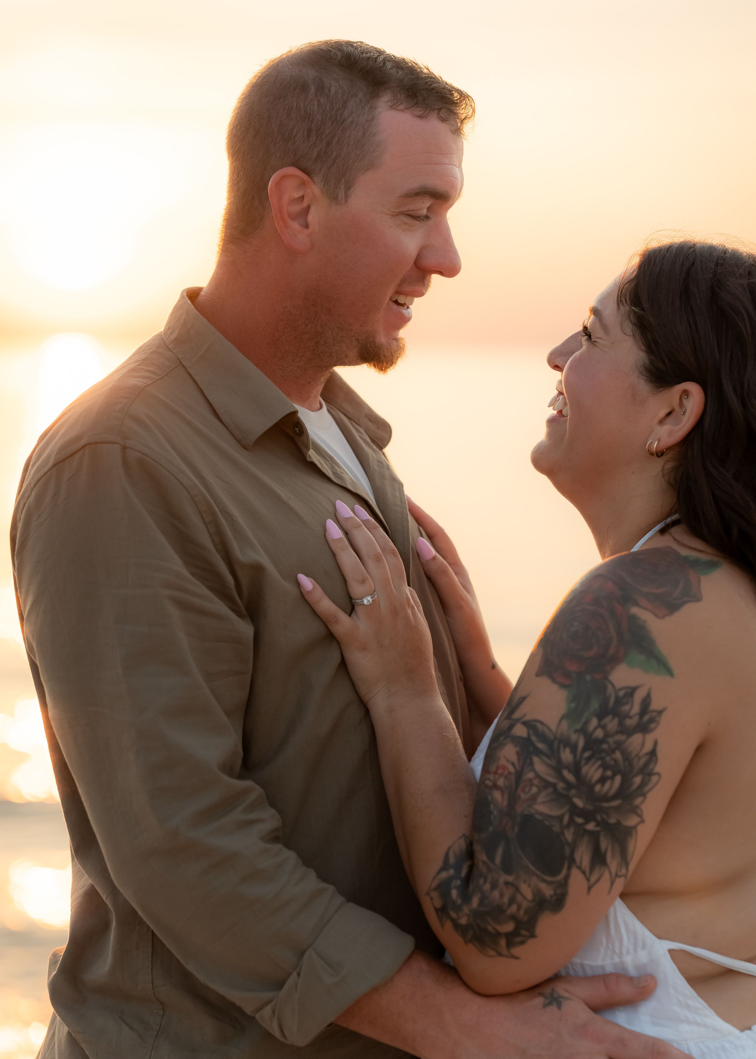 Couple embracing on beach at golden sunset, woman with floral arm tattoo and engagement ring smiling up at man.