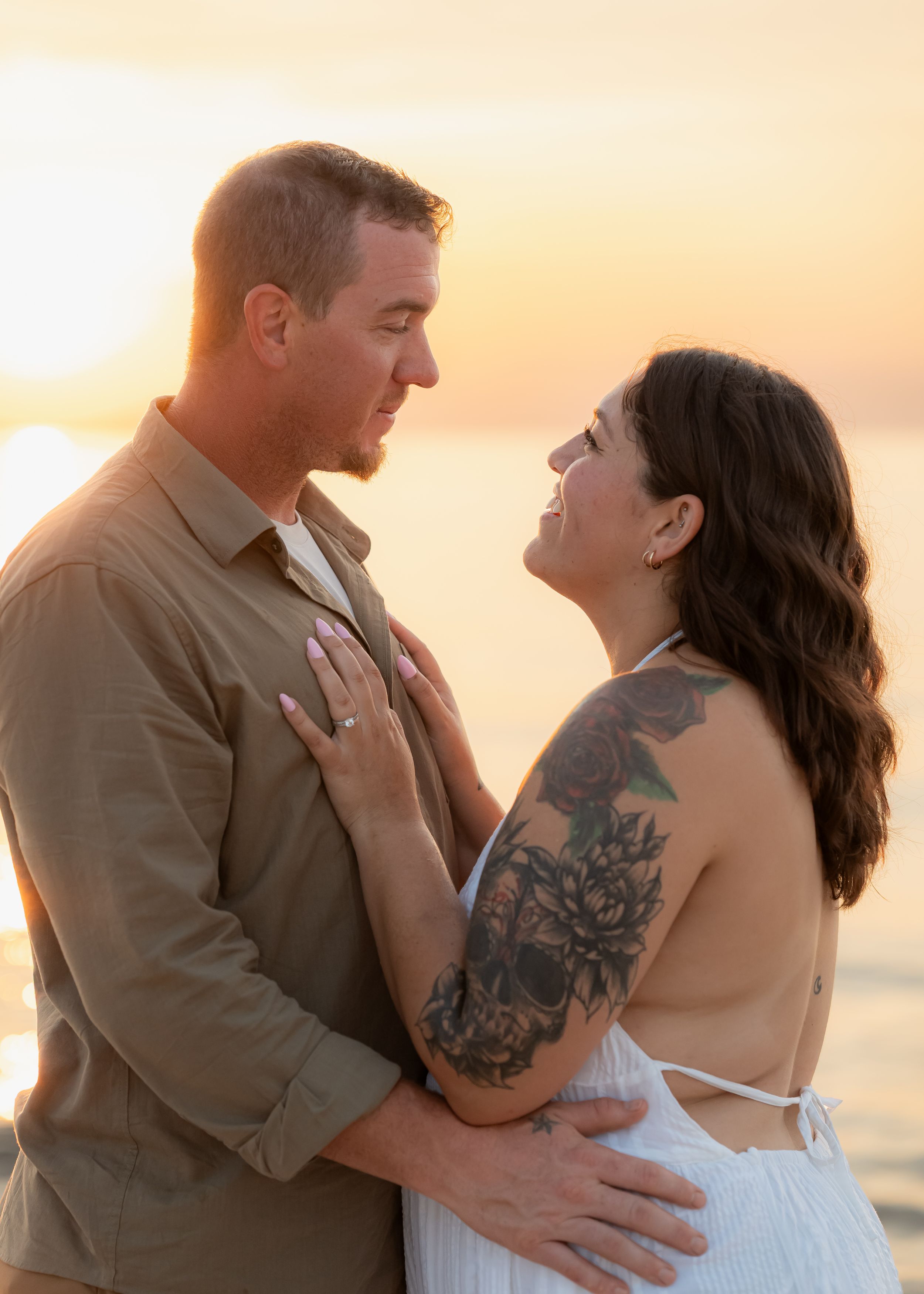 Couple embracing at sunset on the beach, woman with floral arm tattoo in white dress, golden hour backdrop.