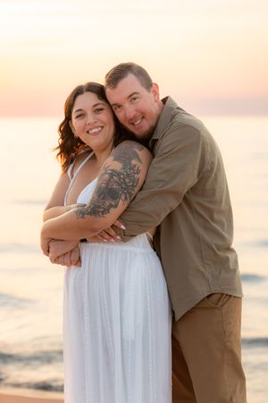 Couple embracing at sunset on the beach, woman in white dress with tattoo, man in olive shirt smiling.