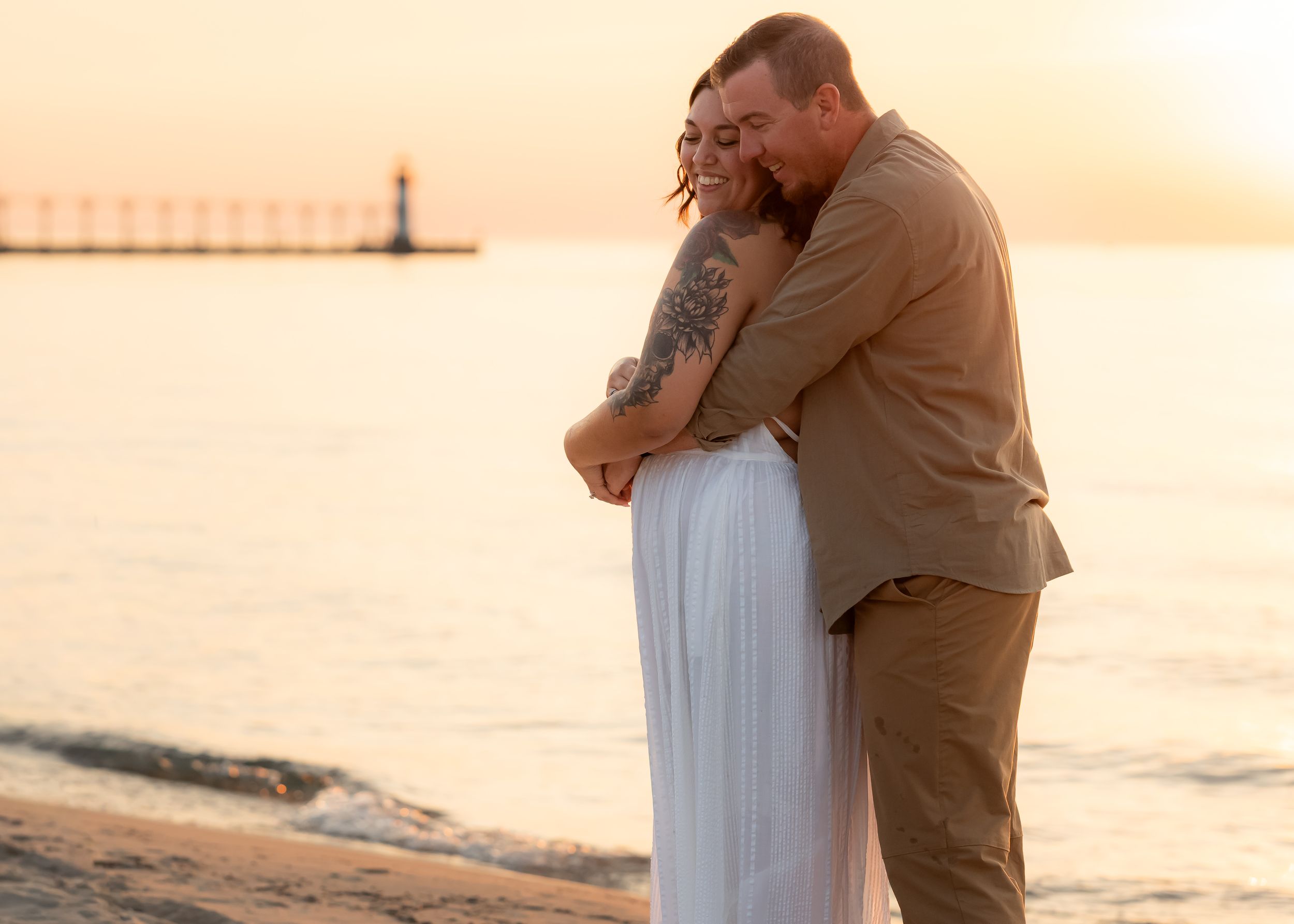 Couple embracing romantically on a beach at sunset near a lighthouse, woman in white dress with tattoo.