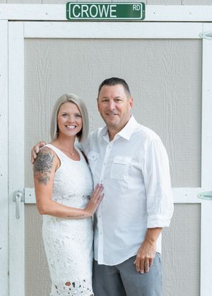 A smiling couple poses together in white outfits beneath a Crowe Road street sign on a sunny day.