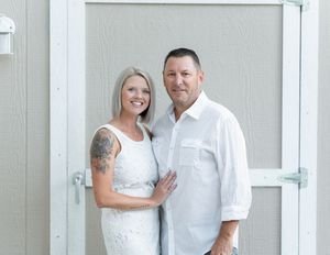 Couple in white outfits smiling together outdoors, woman with tattoo sleeve, man in white button-up shirt.