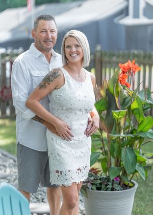 A smiling couple poses outdoors near a potted orange canna lily, the woman in a white lace dress and man in a gray shirt.