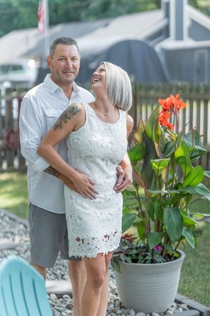 Happy couple embracing outdoors in garden with red canna flowers, man with tattoo hugging woman in white lace dress.