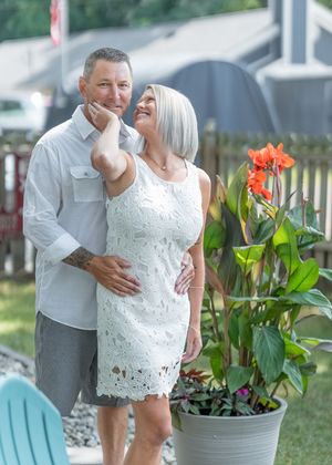 Couple embracing outdoors near tropical plants and orange canna lilies, woman in white lace dress smiling up at tattooed man.