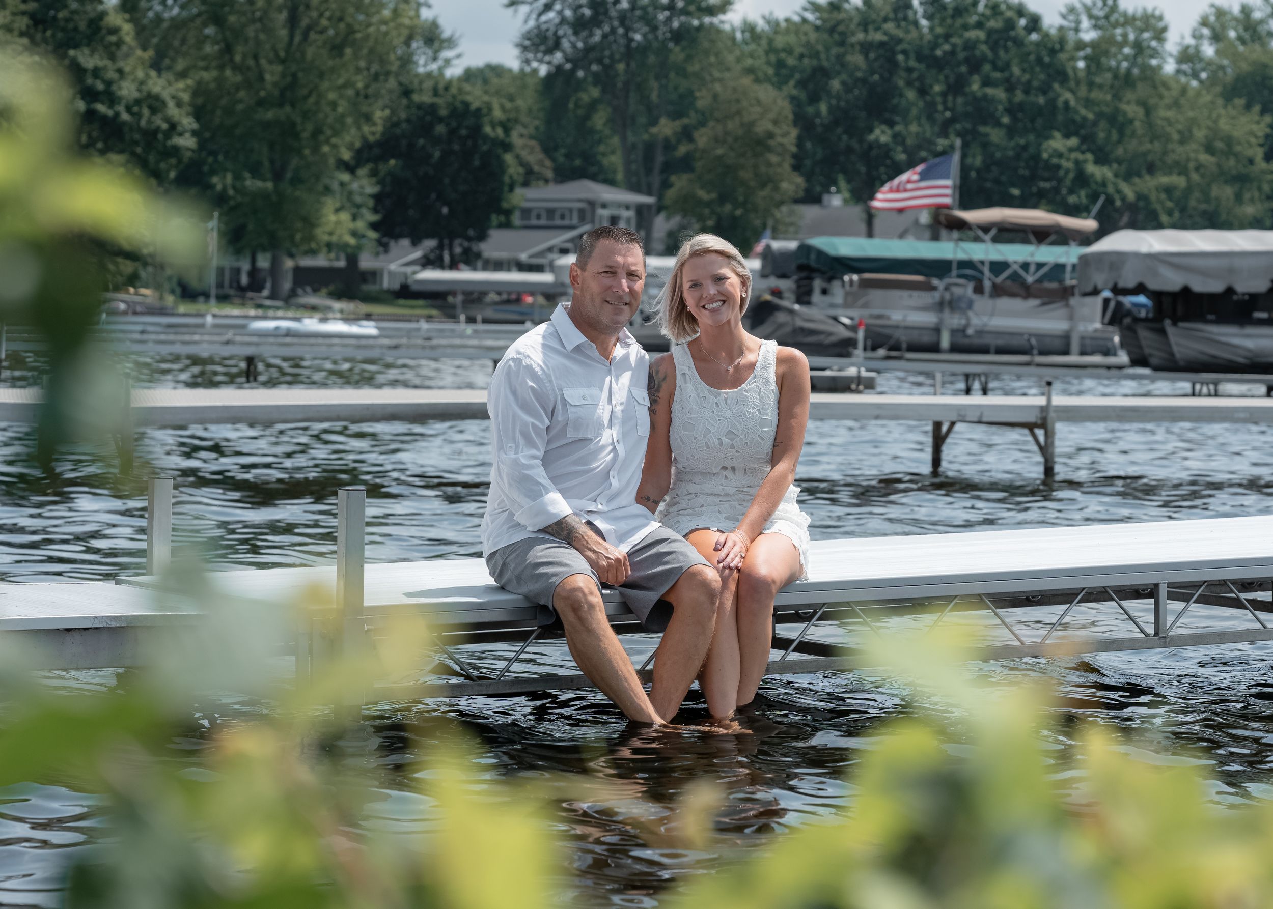 Couple sitting on a dock with feet in the water, smiling, with a scenic lake and trees in the background.