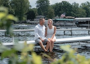 Couple sitting on a dock with feet in the water, smiling at each other, with a lake and boats in the background.