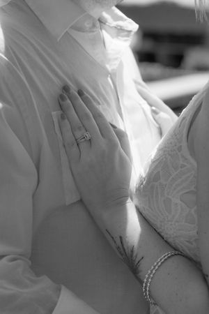 Black and white photo of a bride's hand with engagement ring resting on groom's chest, showing floral arm tattoo.