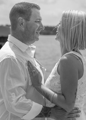 Black and white photo of a smiling couple in wedding attire gazing at each other outdoors near water.
