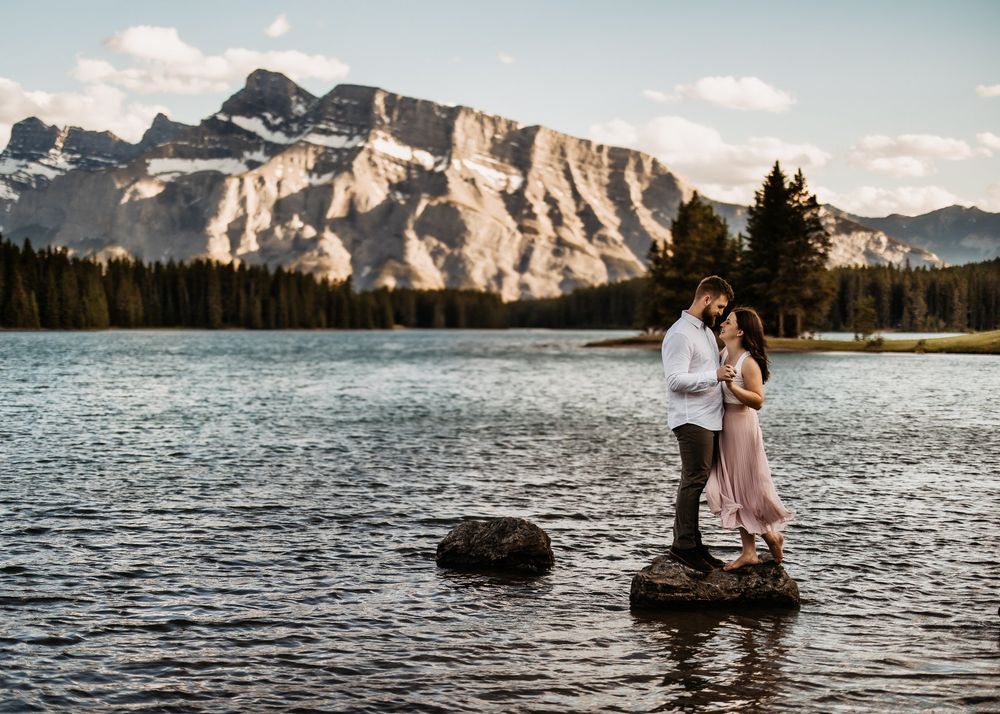 Couple standing on a rock in a turquoise mountain lake below rugged peaks, by a Banff wedding photographer