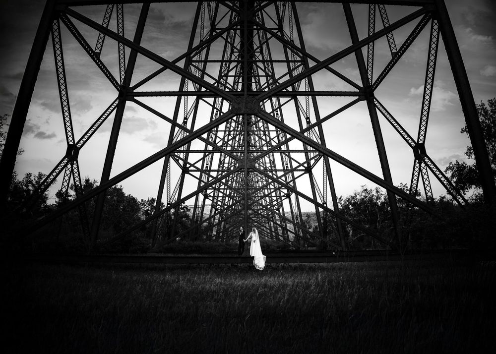 Black and white wedding photo of a bride standing beneath a massive steel bridge