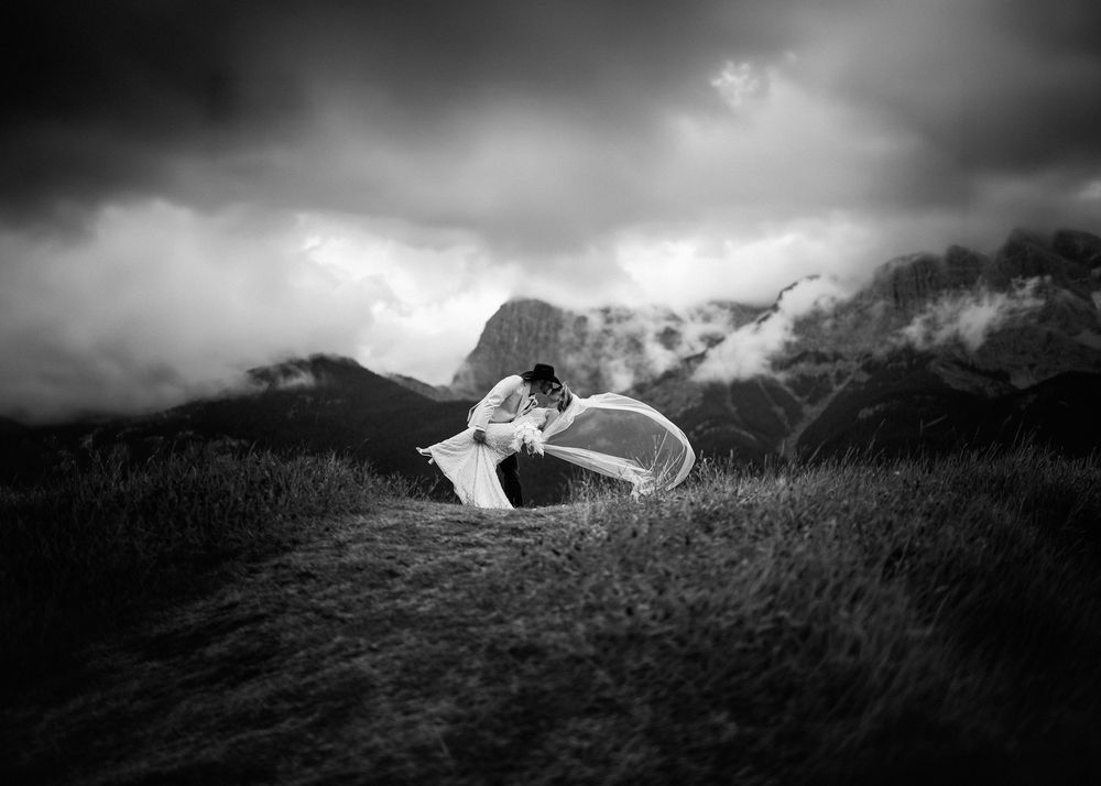 Black and white photo of a groom twirling his bride beneath swirling mountain clouds, by a Canmore wedding photographer