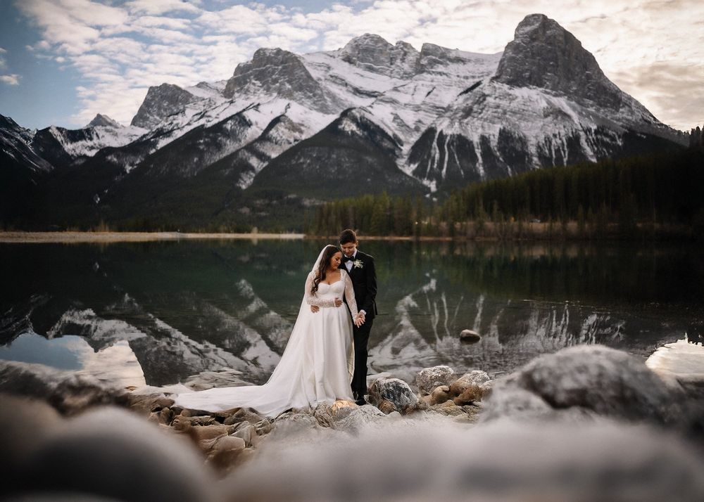 Bride and groom at a reflective lake below snow-capped peaks, by a Canmore wedding photographer