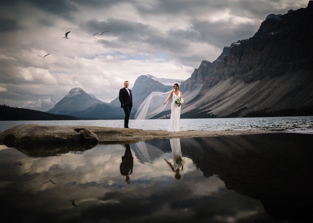 Bride and groom reflected in a still glacial lake beneath towering peaks, by a Lake Louise wedding photographer