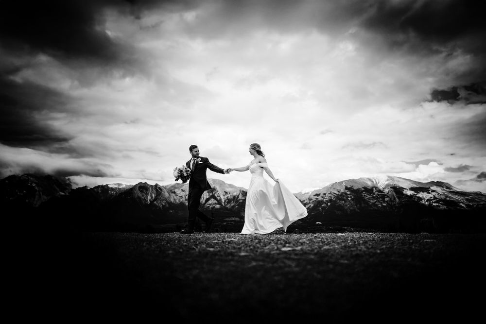 Black and white photo of a bride and groom walking under a dramatic sky with mountains on the horizon