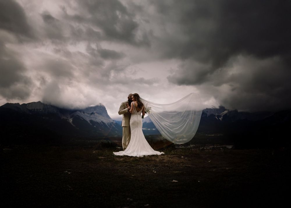 Bride and groom with her veil streaming in the wind beneath storm clouds over the Rocky Mountains