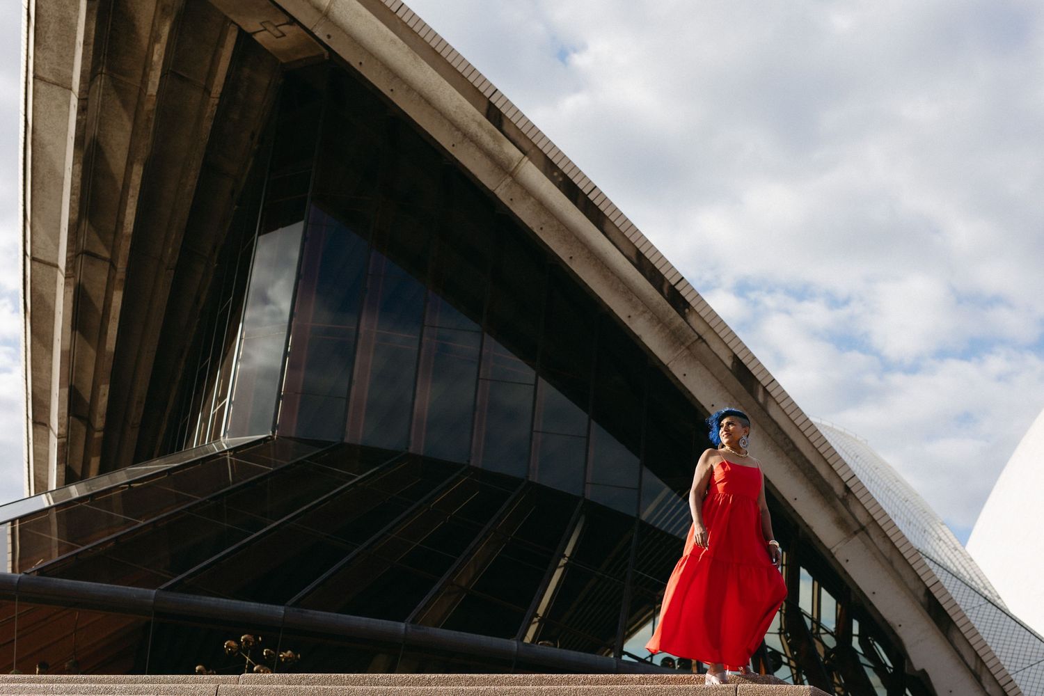 Woman in red dress stands before Sydney Opera House's iconic white shell roof against a cloudy blue sky.