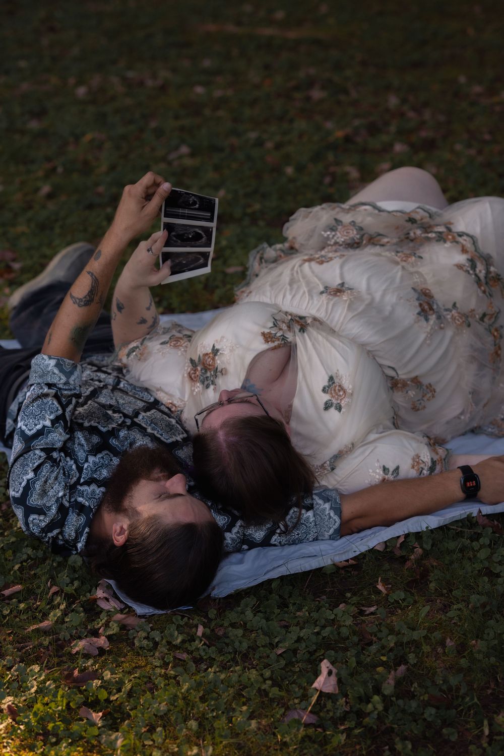 Couple lying on grass looking at ultrasound photos, woman in floral dress, maternity announcement photo.