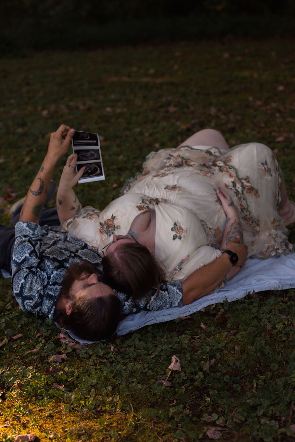 Couple lying on grass holding ultrasound photo, woman in floral dress with baby bump, romantic maternity announcement outdoors.