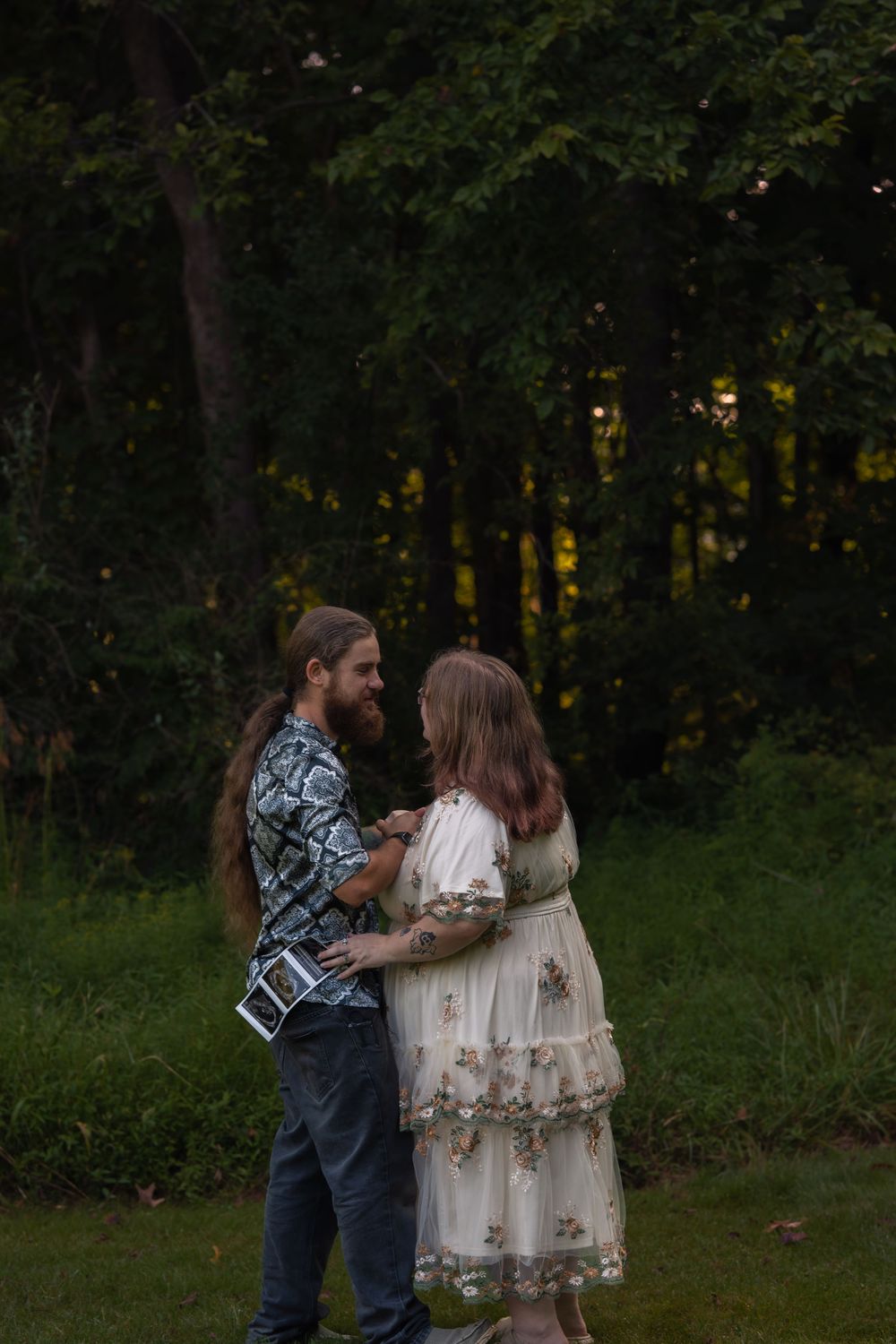 A bearded man with long hair holds a sonogram while embracing a woman in a floral dress outdoors at dusk.