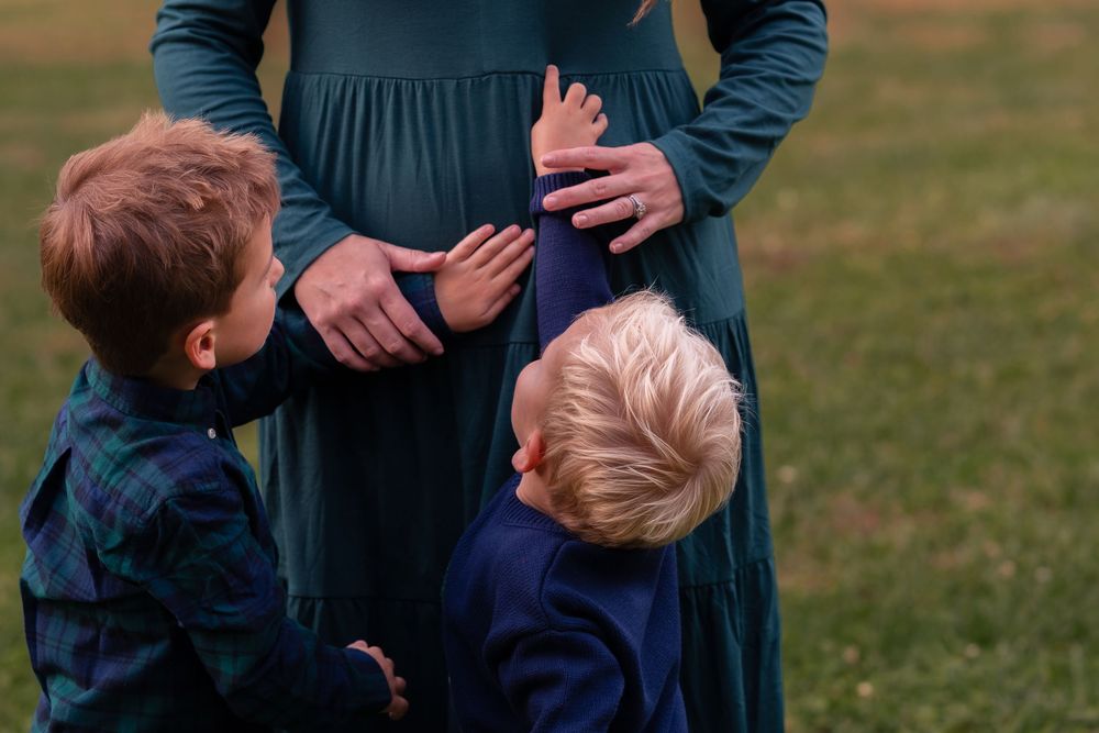 Two young boys touching their pregnant mother's baby bump outdoors on green grass.
