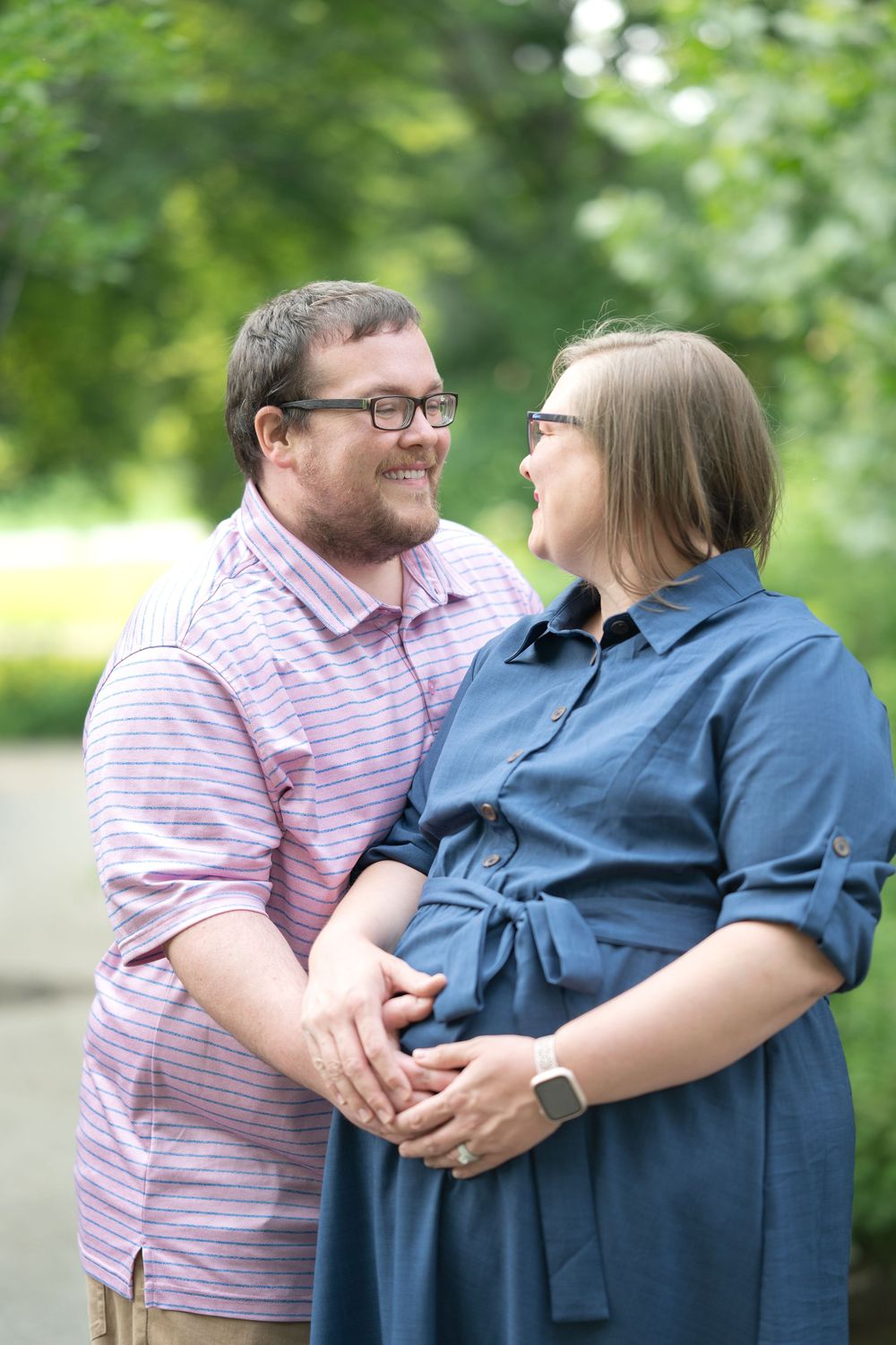 Couple holding hands outdoors, smiling at each other, woman in blue dress appears pregnant, man in pink striped shirt.