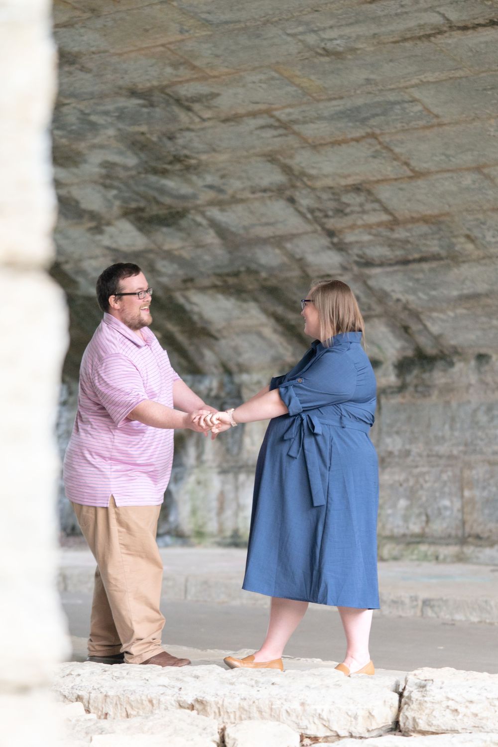 Couple holding hands and smiling near a stone wall outdoors, man in pink shirt and woman in blue dress.