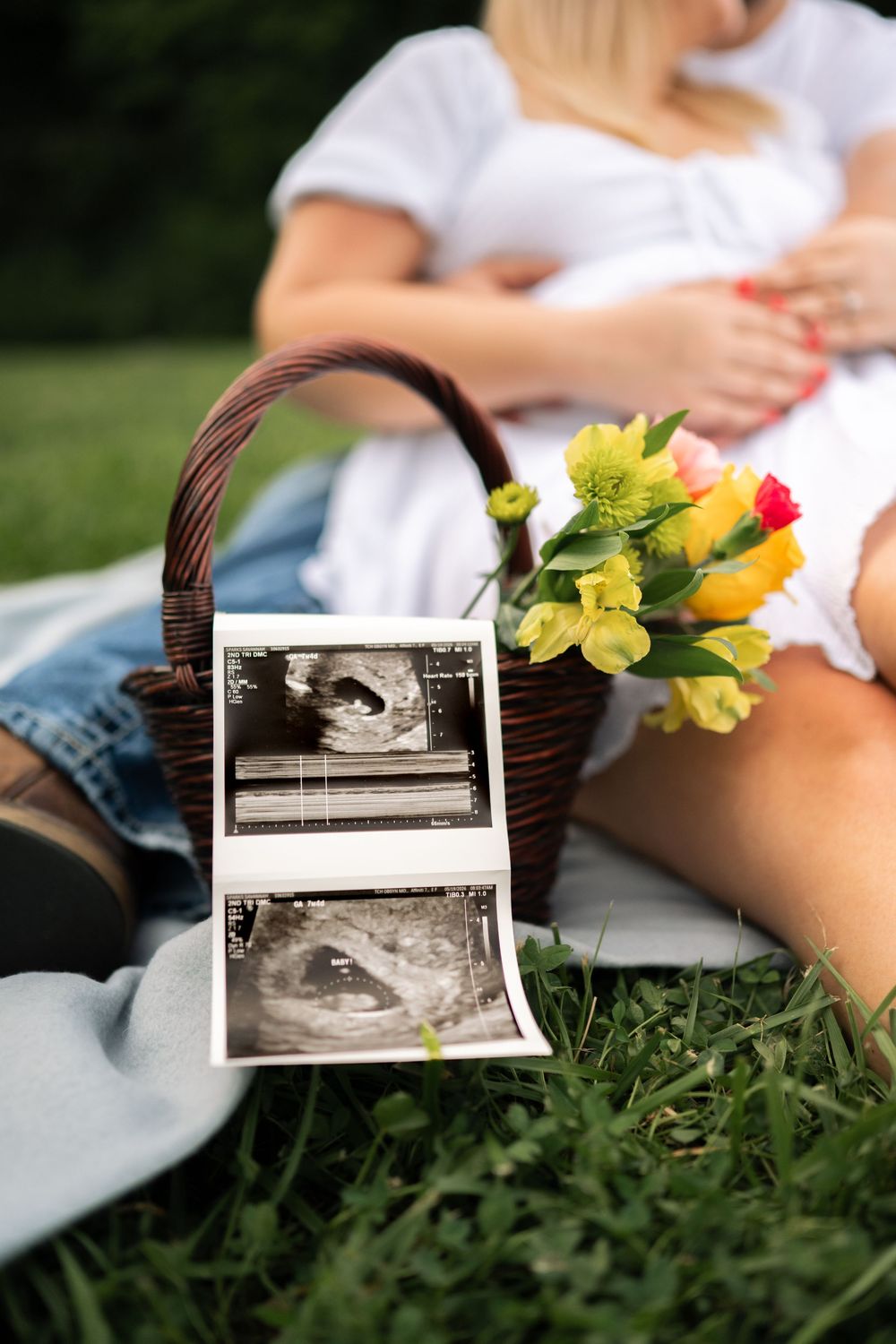 Pregnant couple sitting on grass with ultrasound photos and a wicker basket of colorful tulips during an outdoor picnic.