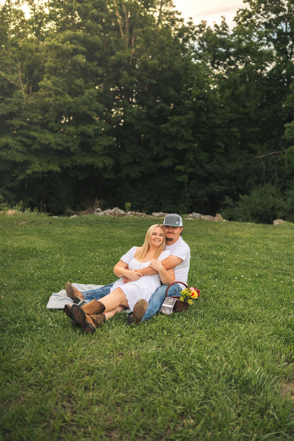 Couple sitting together on green grass in front of trees, woman in white dress and boots, man in cap embracing her.