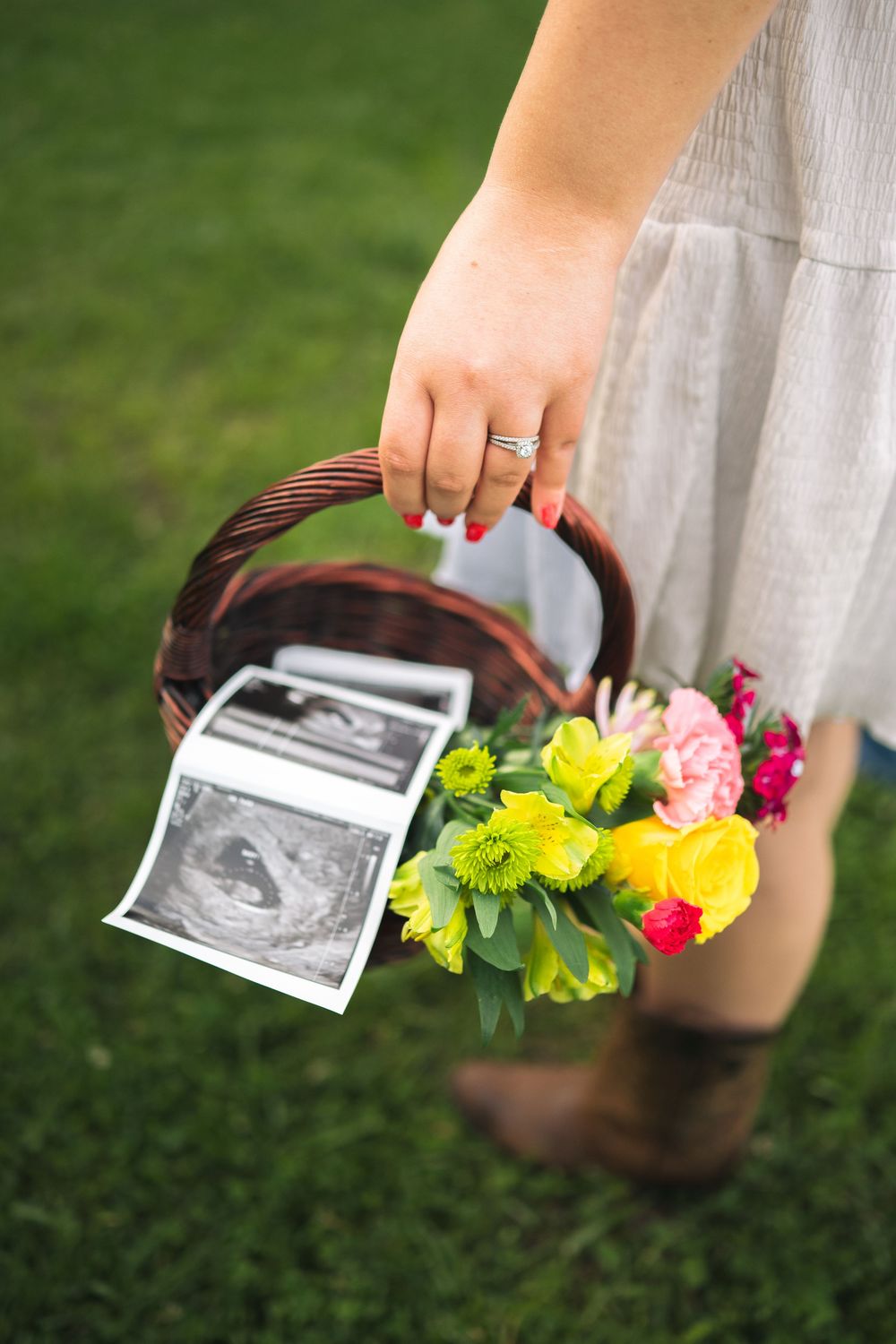 Woman holding a wicker basket with ultrasound photos and colorful flowers on green grass, wearing a ring.