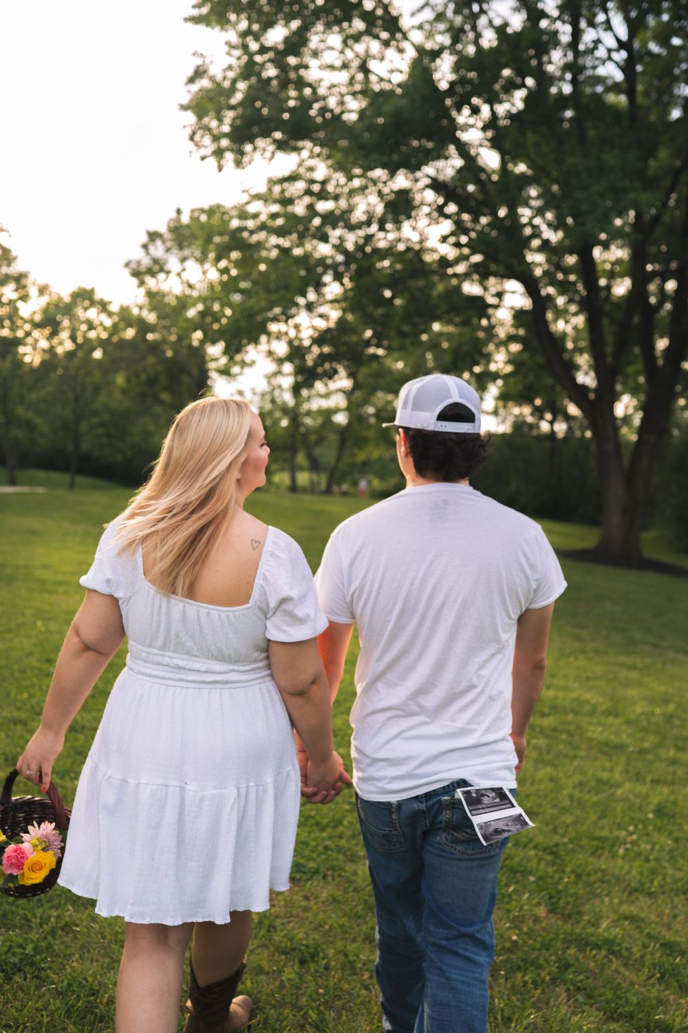 A couple walks hand-in-hand through a lush green park at sunset, woman in white dress holding flowers, man in white tee and cap.