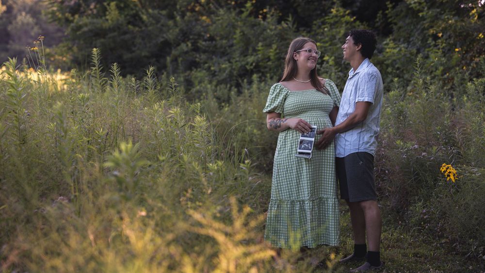 Pregnant couple standing in a lush green field, woman in mint dress holding ultrasound photo, sharing a tender moment.