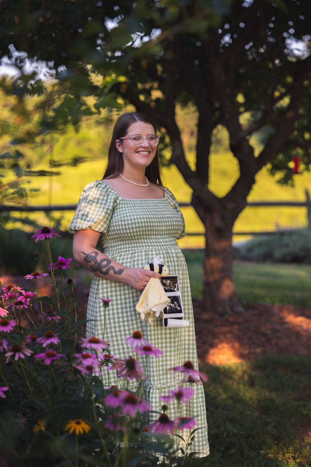 Pregnant woman in green gingham dress holding ultrasound photo, standing among pink flowers in a sunlit garden.