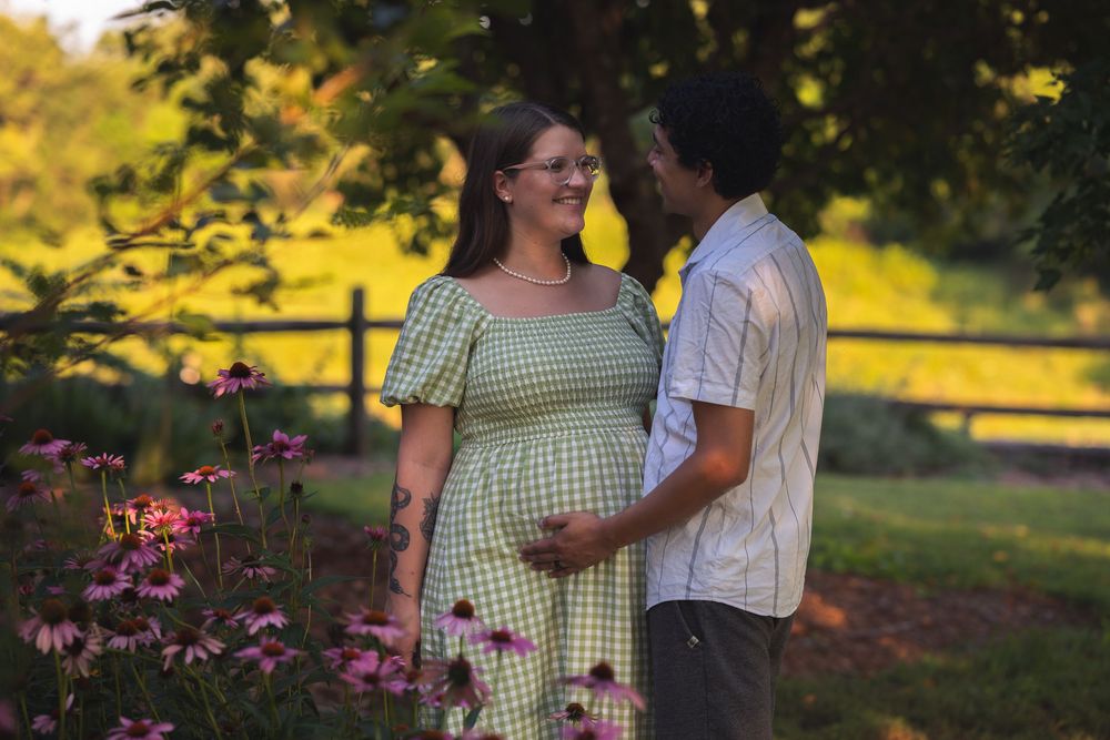 Pregnant couple embracing in a lush garden surrounded by pink flowers during golden hour maternity photoshoot.