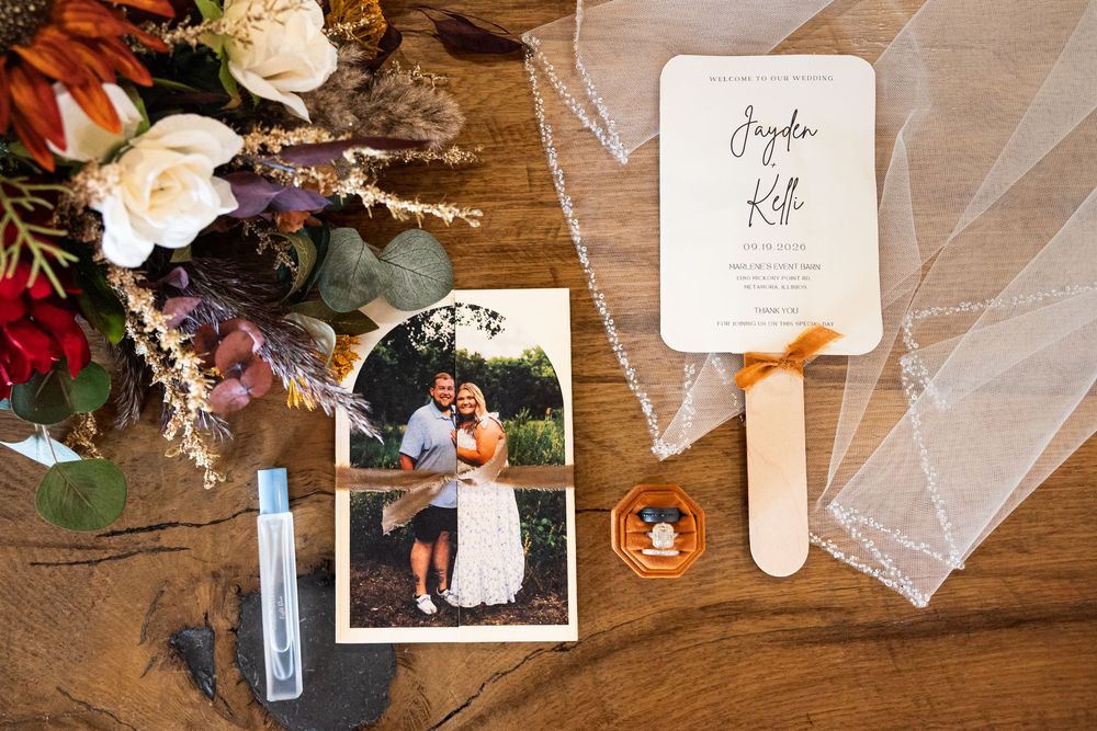 Wedding flat lay with invitation, couple photo, fan program, veil, and floral bouquet on rustic wood table.