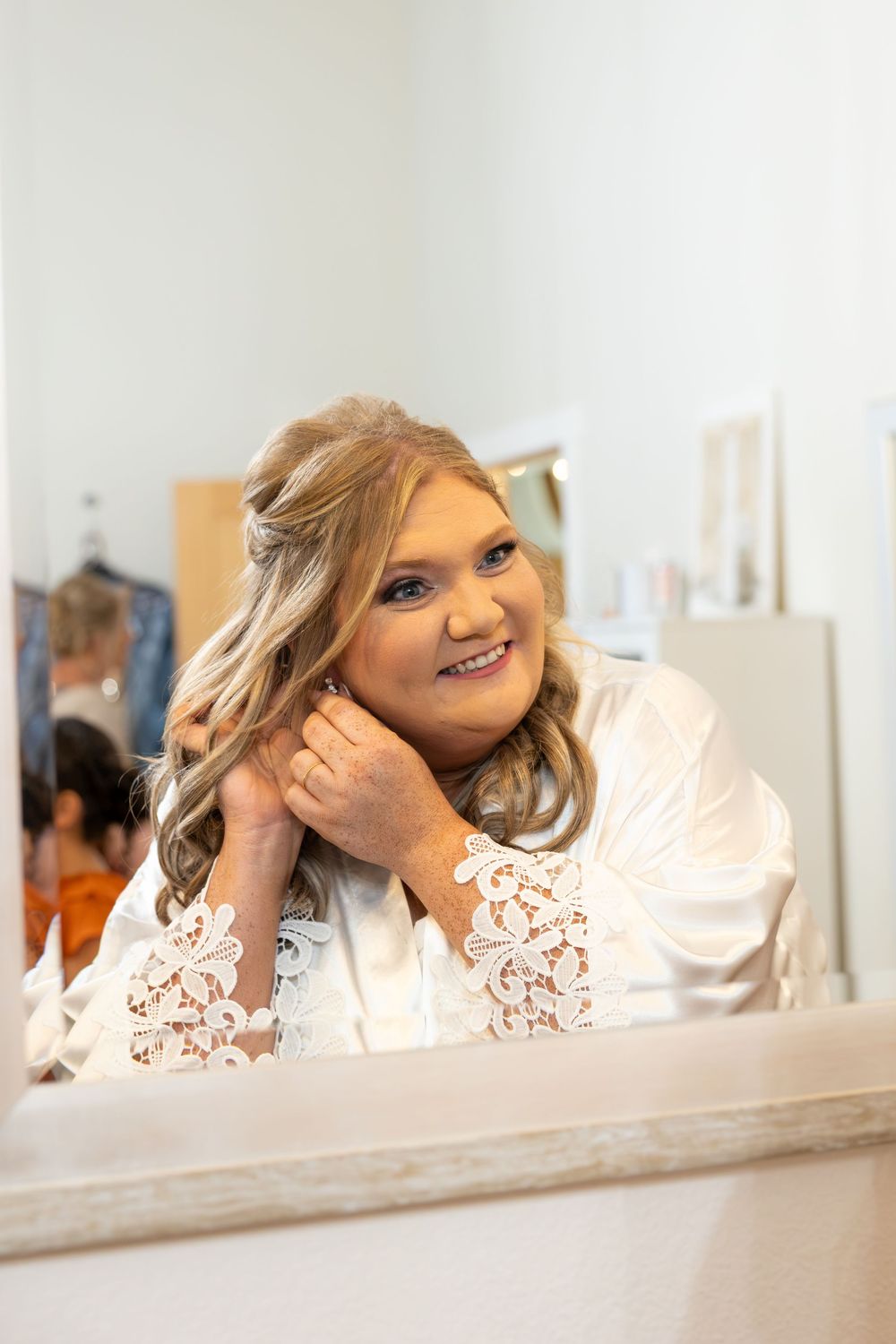 A smiling blonde bride in a white lace robe adjusts her earring while looking in a mirror.
